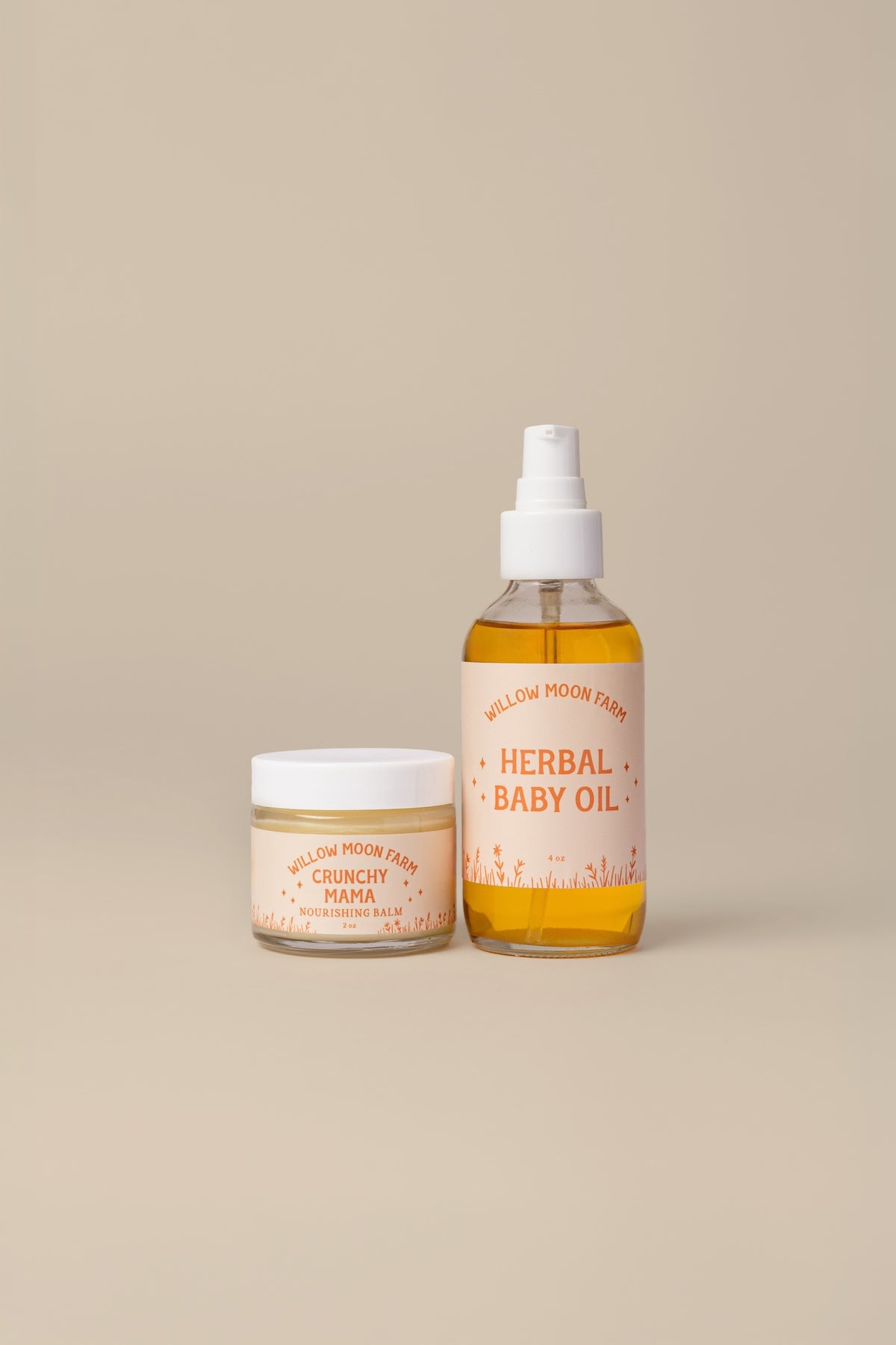 Two products from Willow Moon Farm displayed on a solid beige background: a clear spray bottle of Herbal Baby Oil with amber liquid and a white cap, and a small glass jar of Crunchy Mama Balm with a white lid.