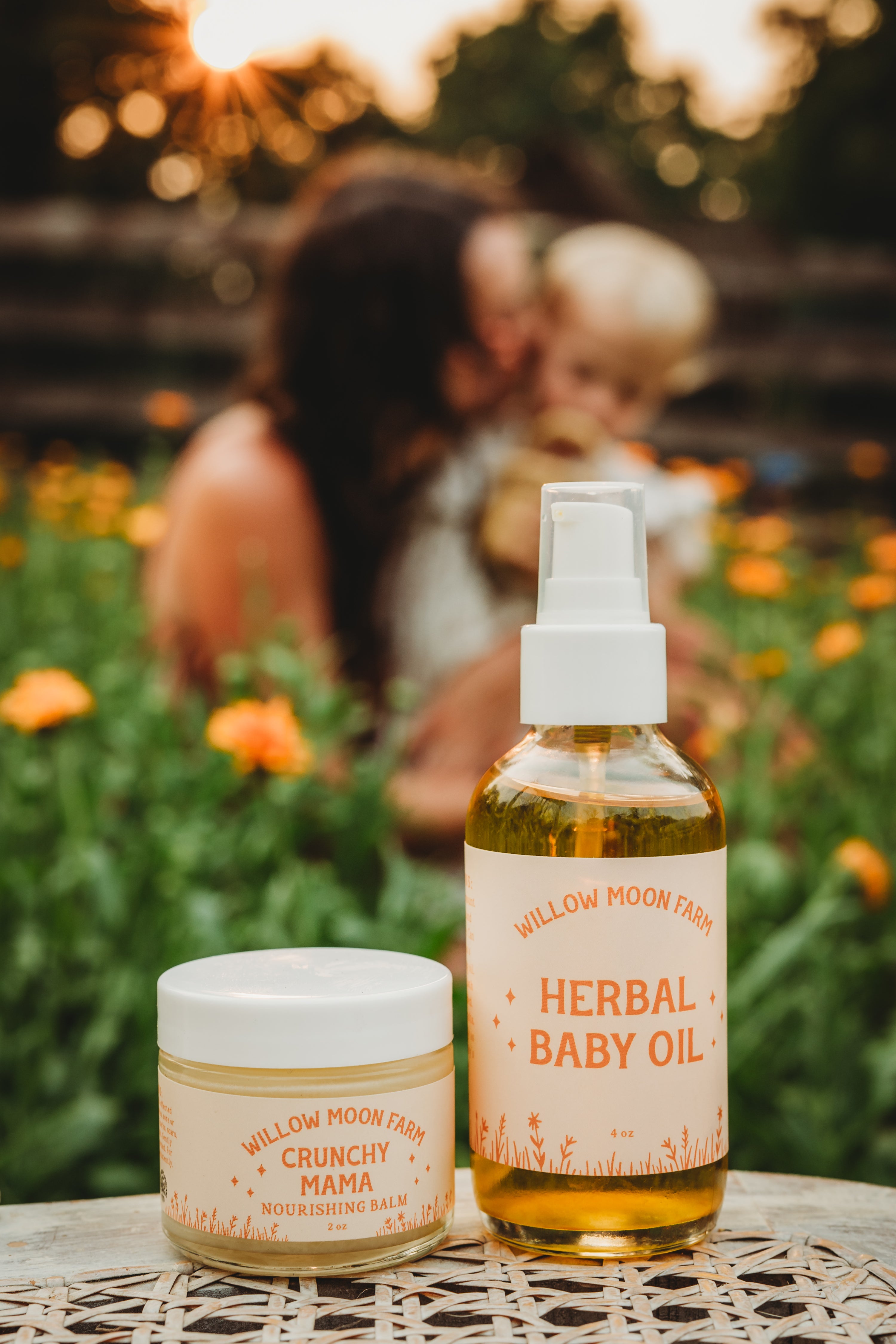Two Willow Moon Farm products displayed on a table in a garden setting: a bottle of Herbal Baby Oil and a jar of Crunchy Mama Moisturizing Balm, with a blurred mother and child in the background.